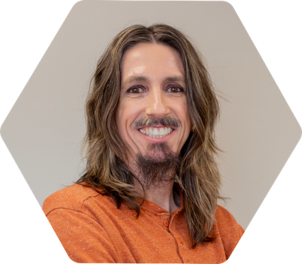 Brian Ricks smiling person with long brown hair in a hexagonal frame, against a white background.
