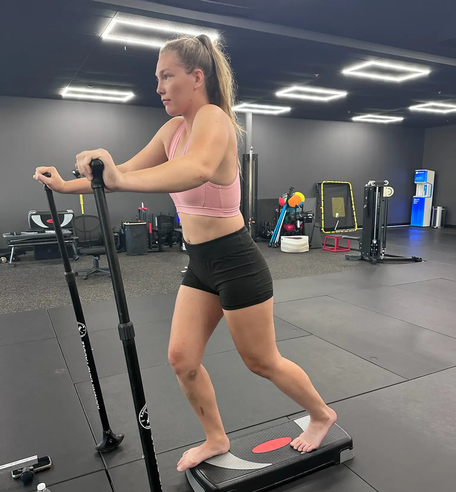 Woman exercising on a stair-stepper in a gym, wearing a pink top and black shorts.