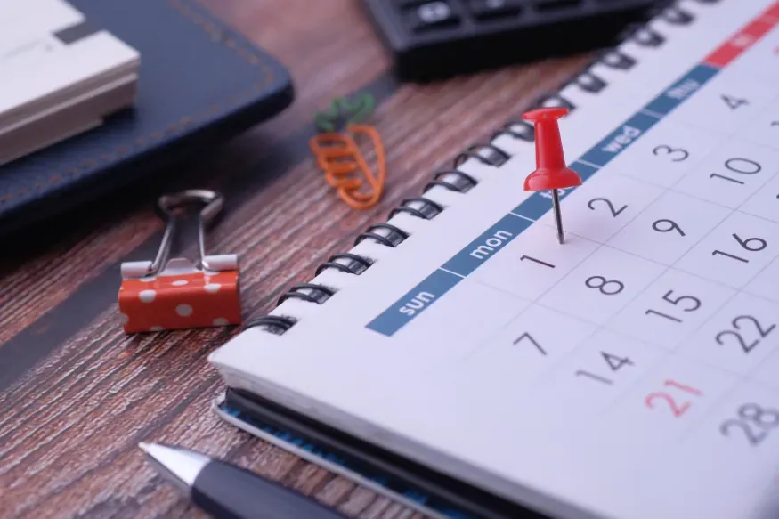 Desk calendar with a red pushpin marking a date on a spiral-bound page next to paper clips.