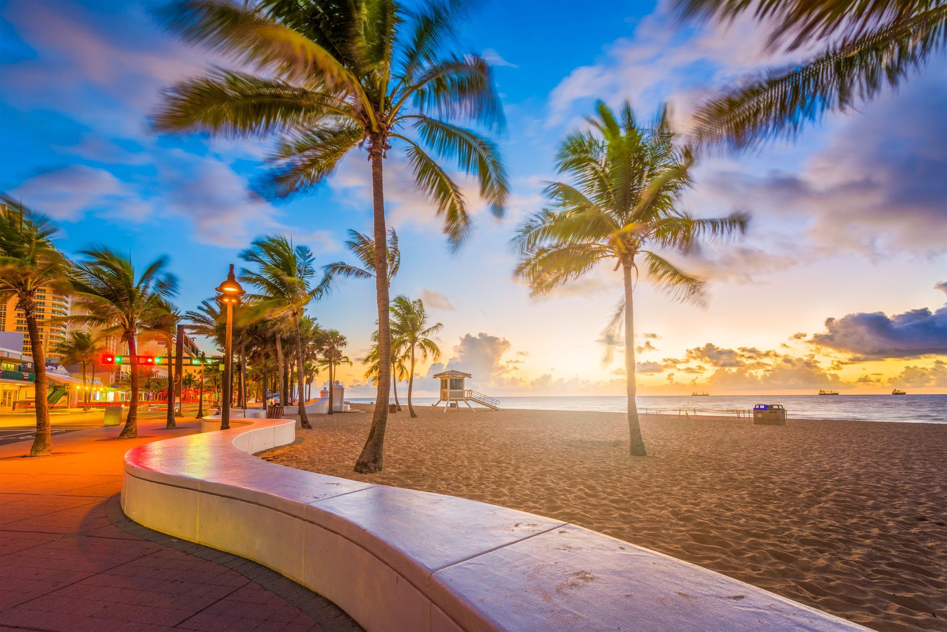 Sunset beach with palm trees, curved walkway, and calm ocean under a colorful sky