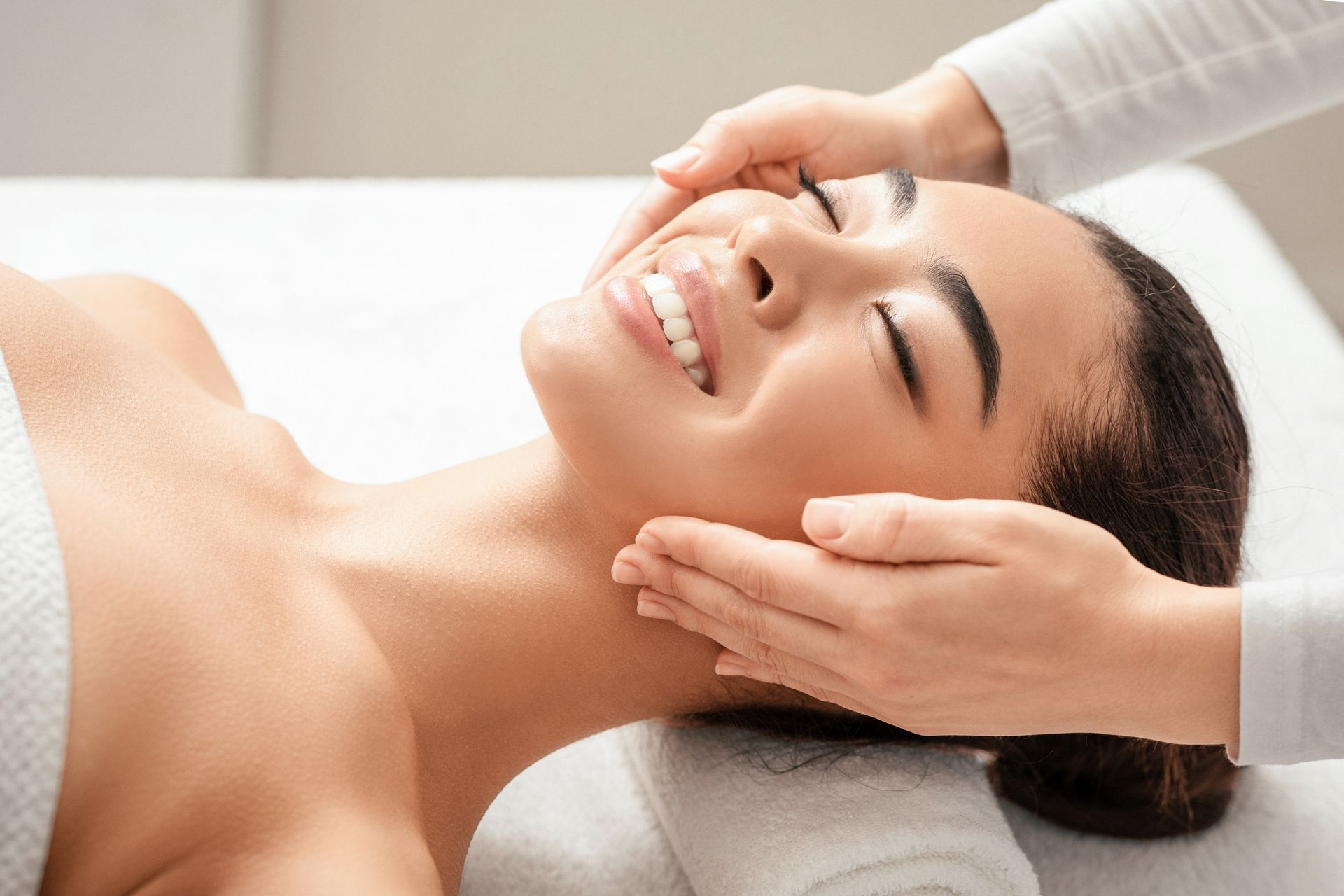 Person receiving facial massage; hands gently touching face, smiling, eyes closed.