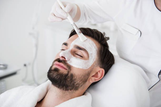 Man receiving facial mask in spa, eyes closed, white mask on face, brush applying product.