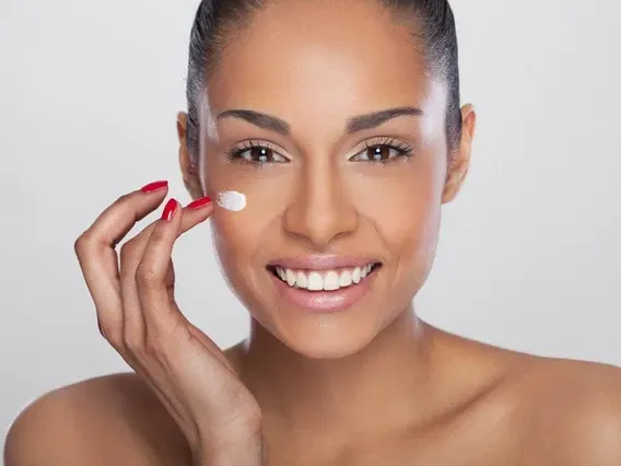 Woman applying cream to her face, smiling. Red nail polish.