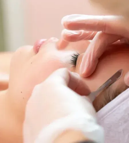 Close-up of a person's face receiving a dermaplaning treatment; gloved hands hold a blade.