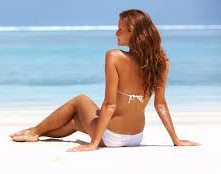 Woman in white bikini sits on beach, looking at ocean.