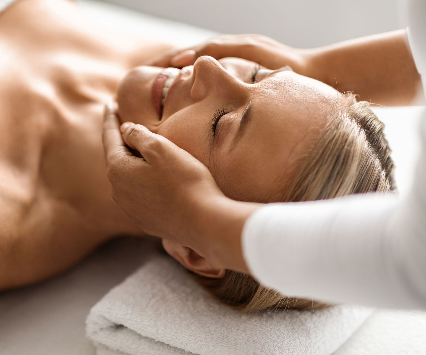 Woman relaxing on a massage table, eyes closed. Red flowers and candle nearby. Bright, neutral setting.