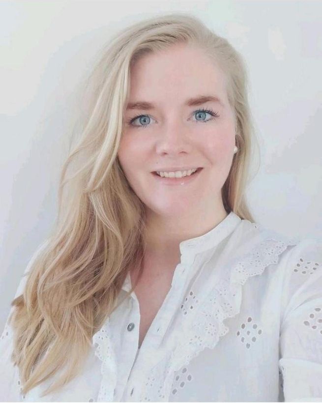 Vrouw met blond haar lacht, draagt ​​een witte blouse en staat voor een lichte achtergrond.