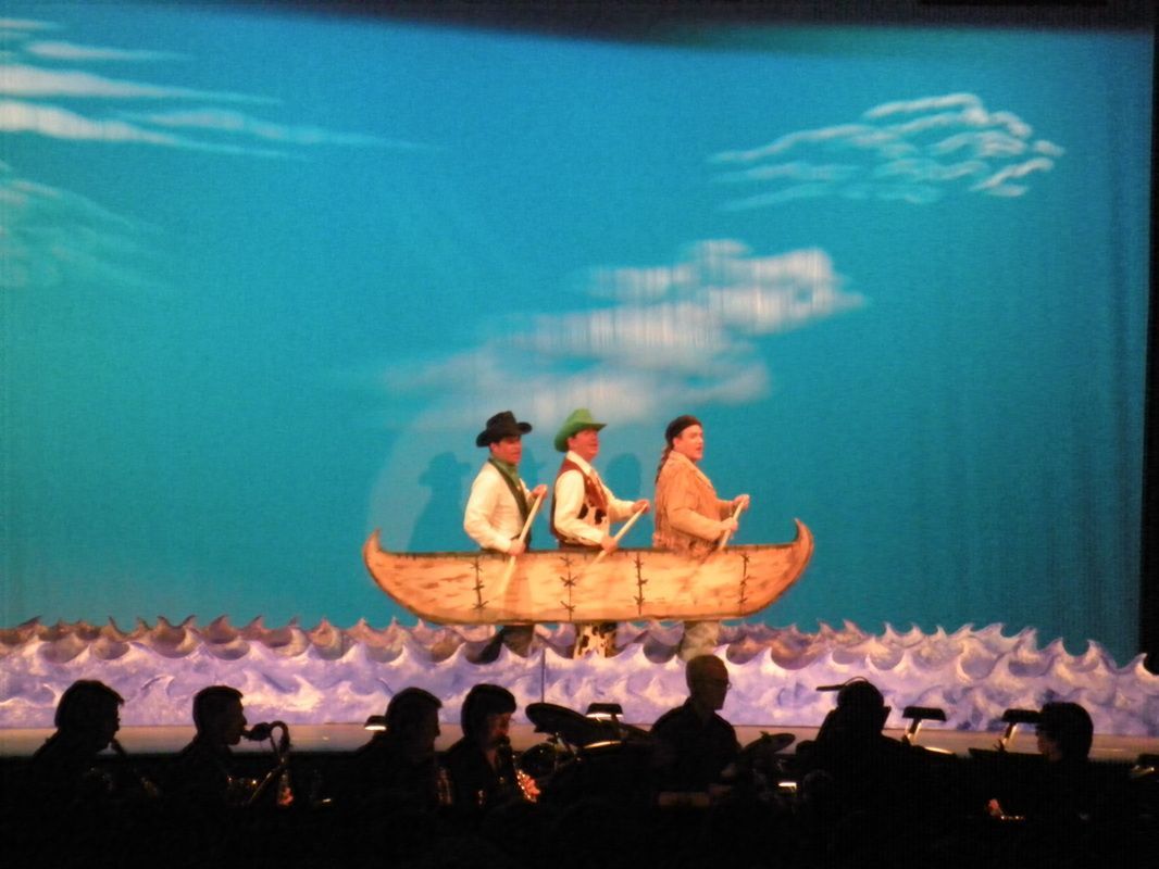 Three people in a wooden canoe on a blue, painted ocean, with a cloudy sky backdrop. A crew of people are seated in the foreground.