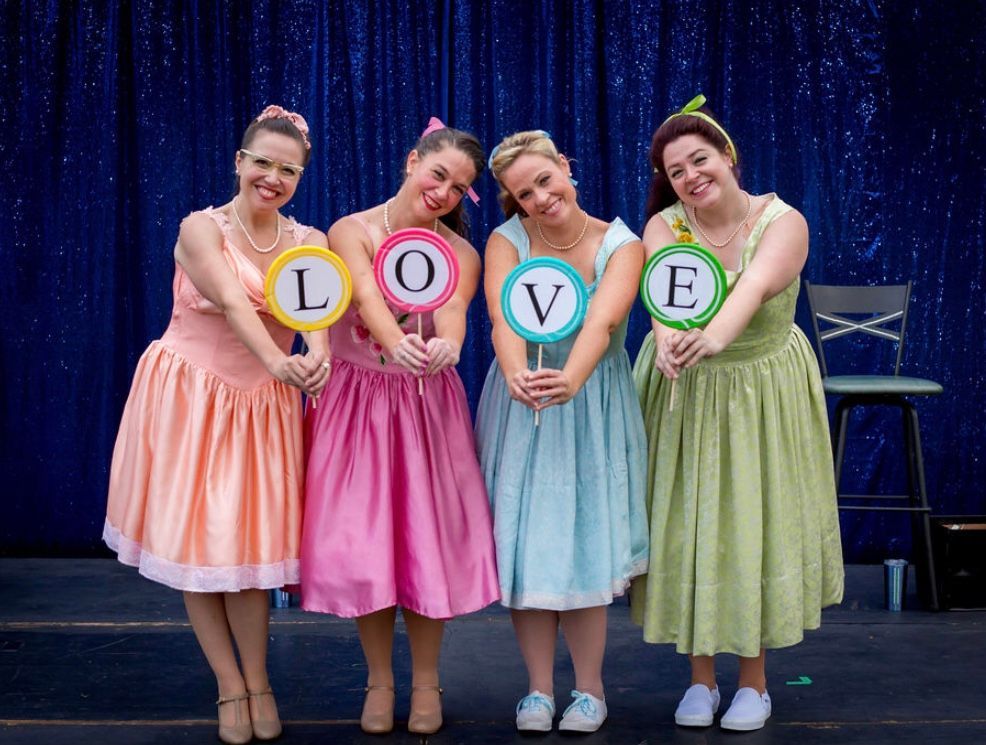 Four women in pastel dresses hold up letters that spell