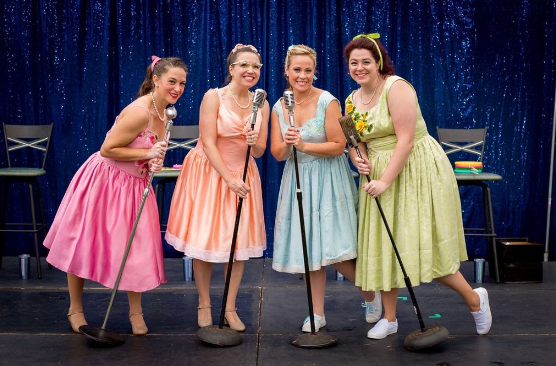 Four women in 1950s dresses sing into microphones on a stage. They are smiling and performing in front of a blue curtain.