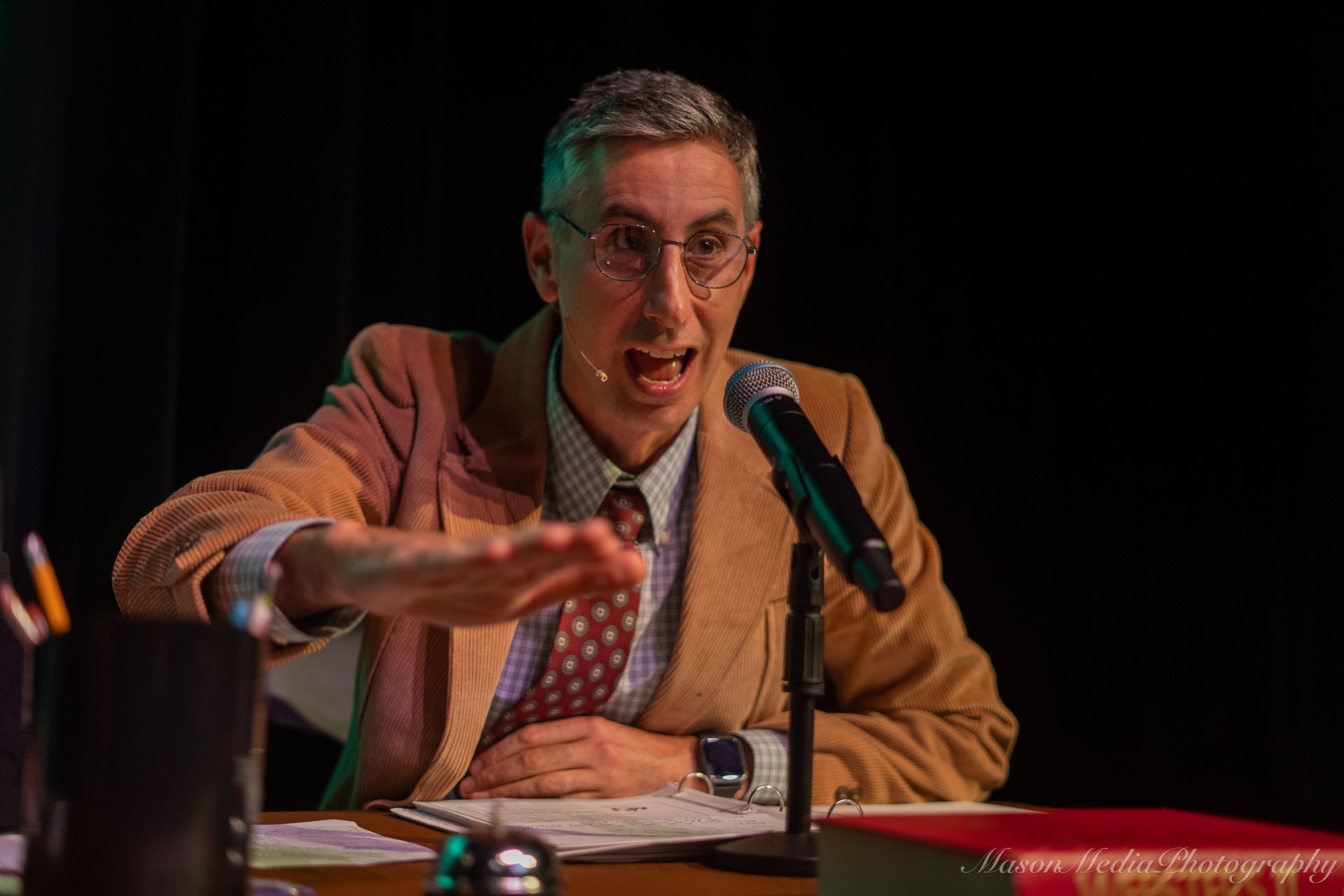 A man in a corduroy jacket speaks into a microphone, gesturing with his hand. He's sitting at a table, with a pen holder and a book.