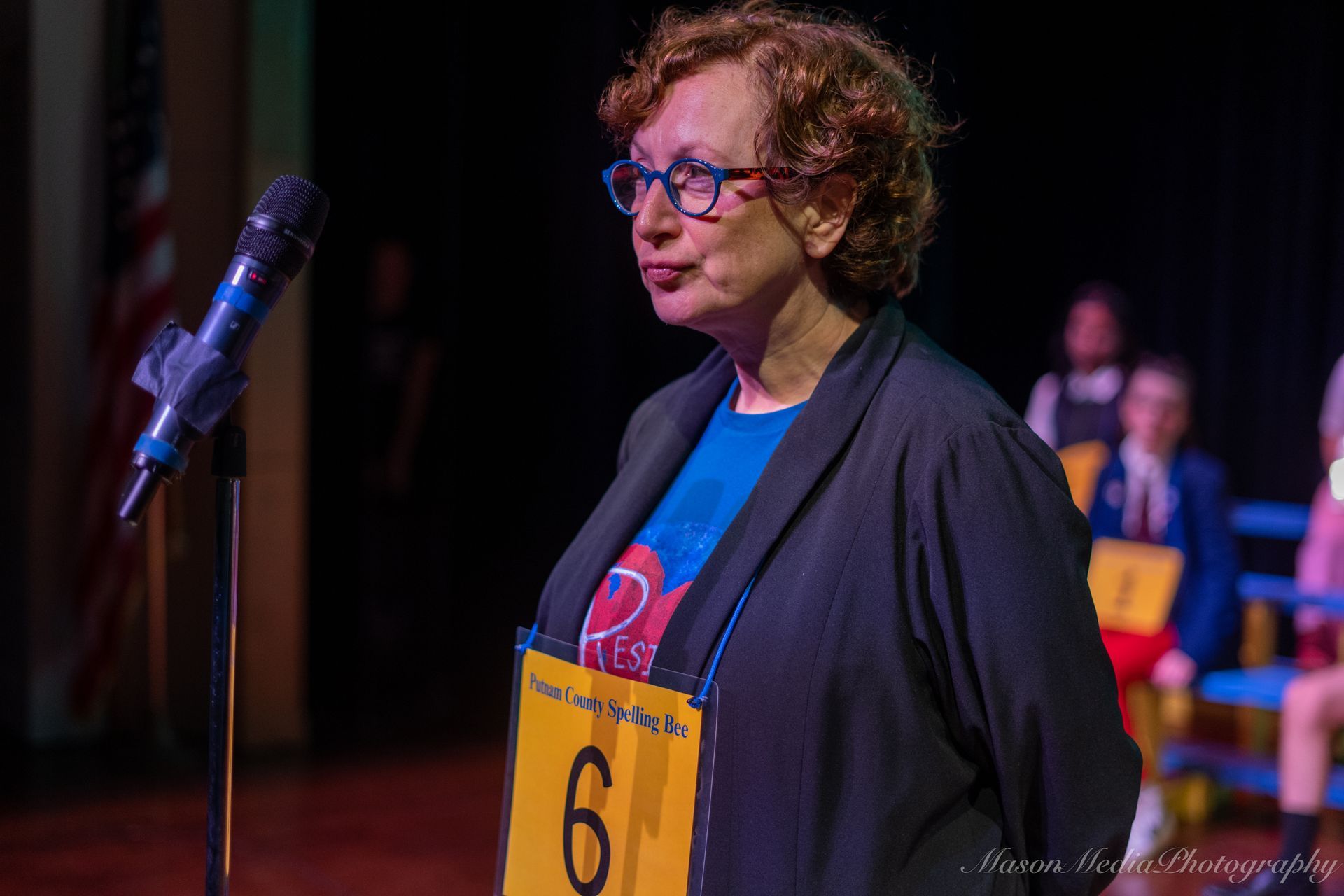 A woman with glasses at a spelling bee, wearing a blue shirt, speaking into a microphone. Stage with contestants in background.
