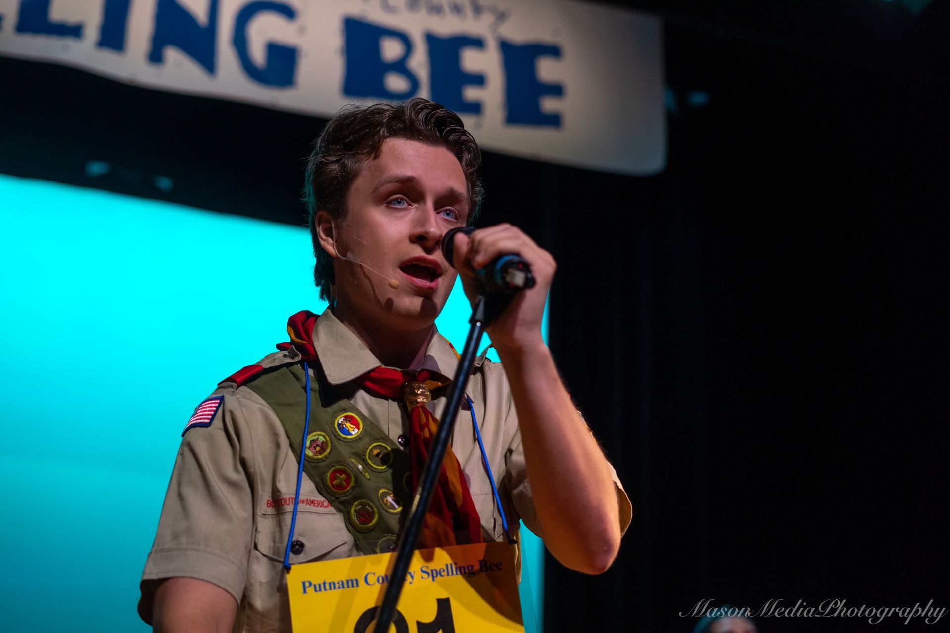 Man in Boy Scout uniform at spelling bee, speaking into microphone. He's on stage with a concerned expression.