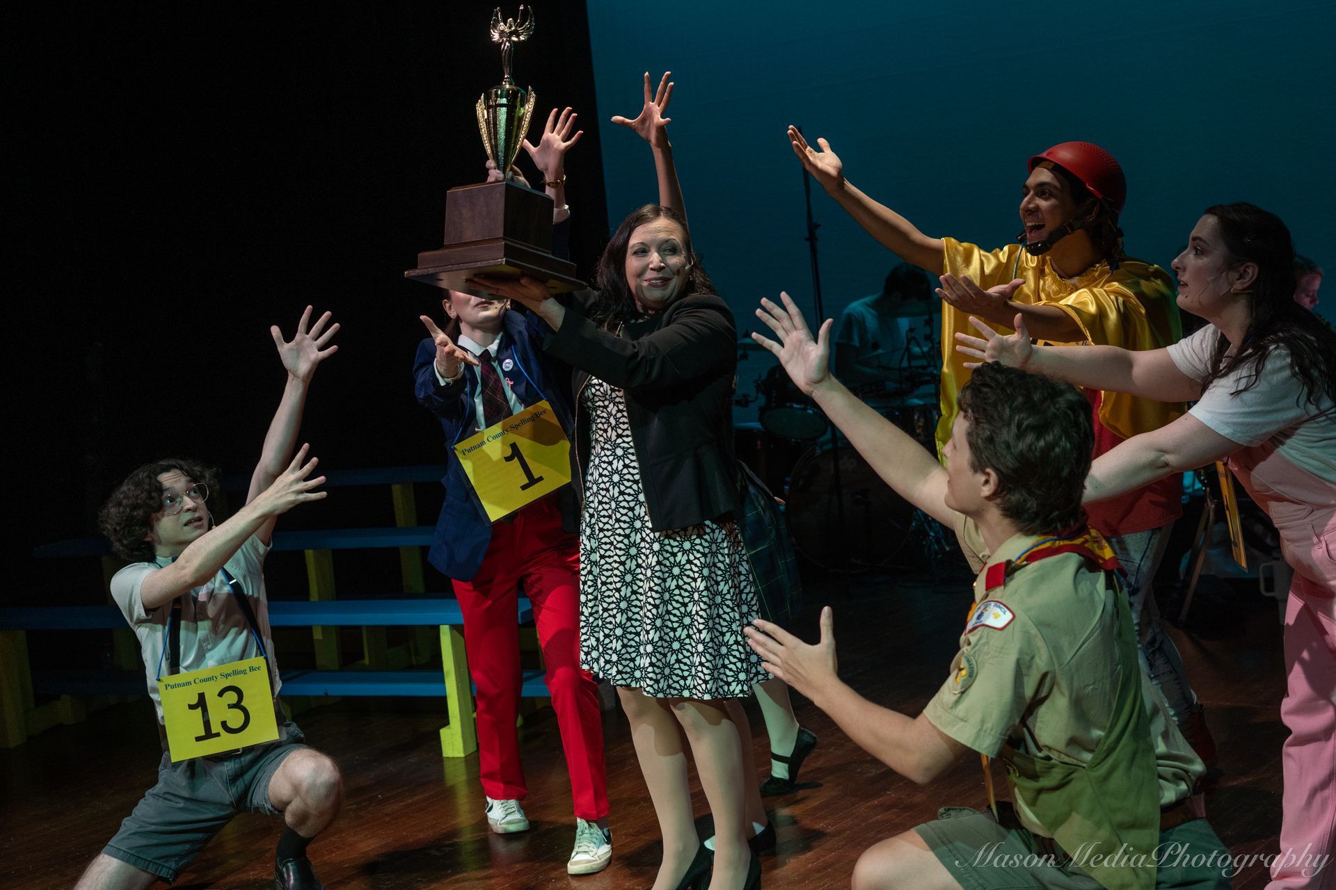 A cast of characters celebrate onstage, holding a trophy. The group has diverse clothing and ages, with excited expressions, set against a dark backdrop.