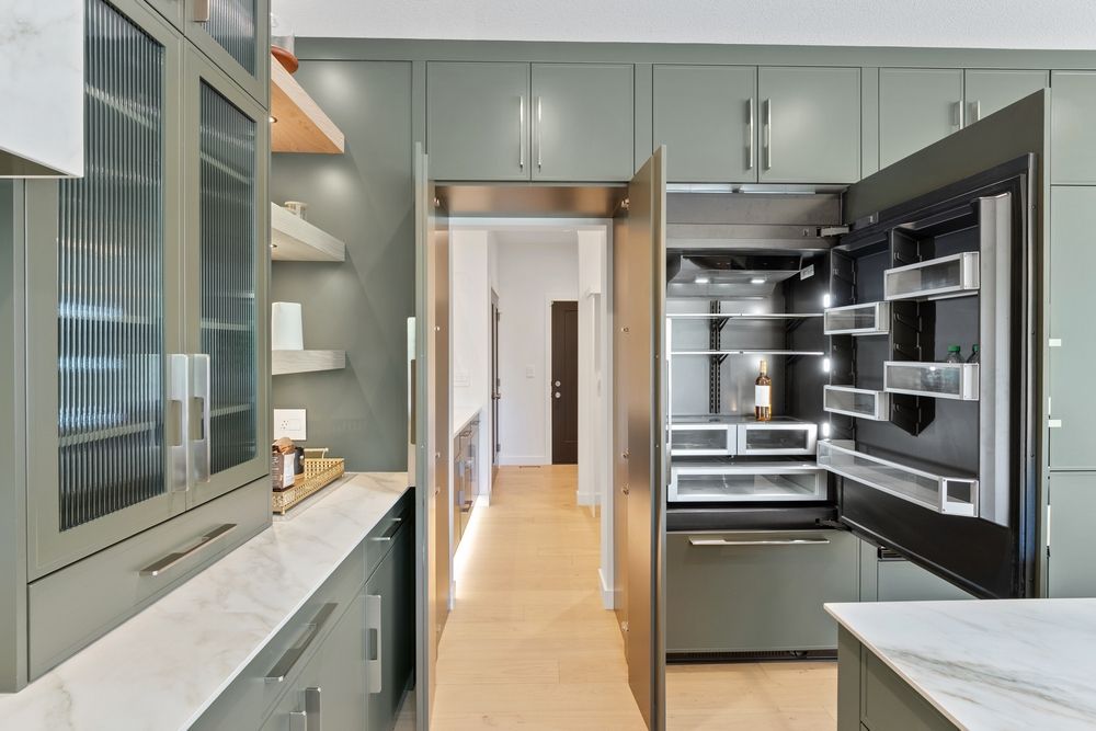 Sage green kitchen with open refrigerator door, light wood floors, and marble countertops.