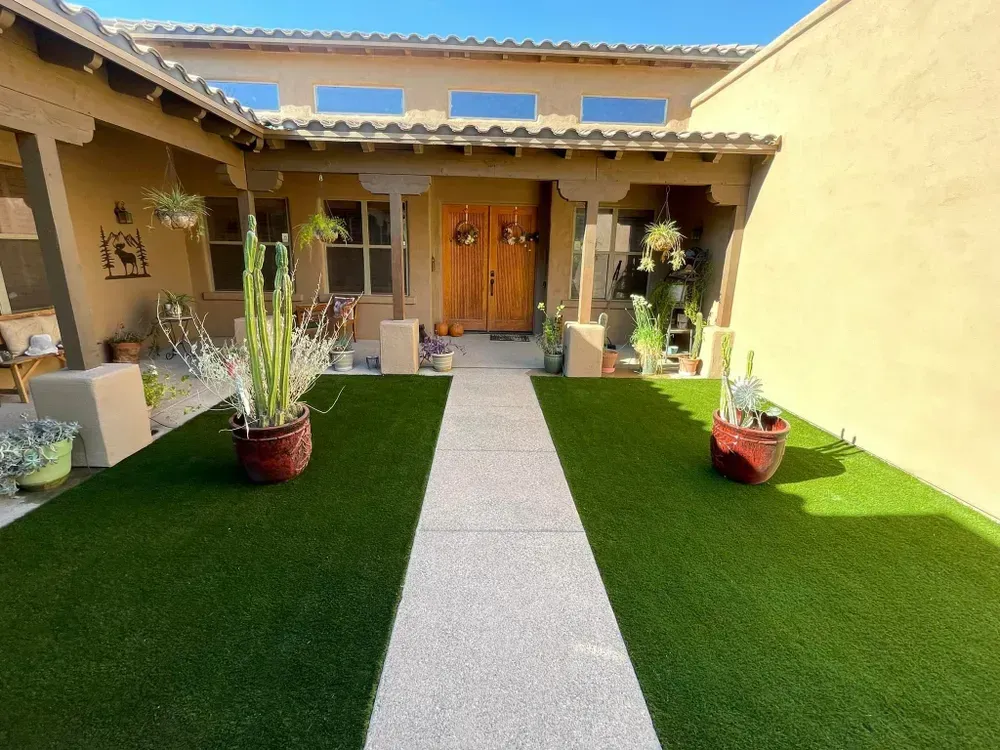 A stucco-style home with a walkway lined by artificial turf and potted cacti, leading to a wooden double door entrance.