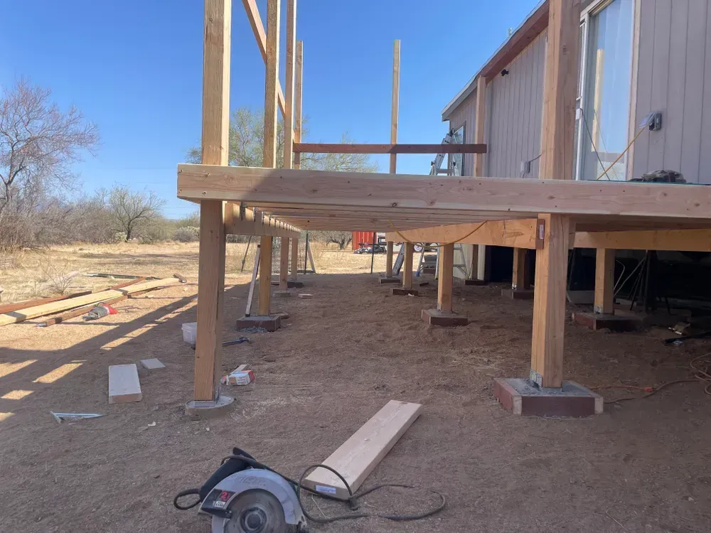 A deck under construction next to a house with exposed wooden beams, a circular saw on the ground, and a clear blue sky.