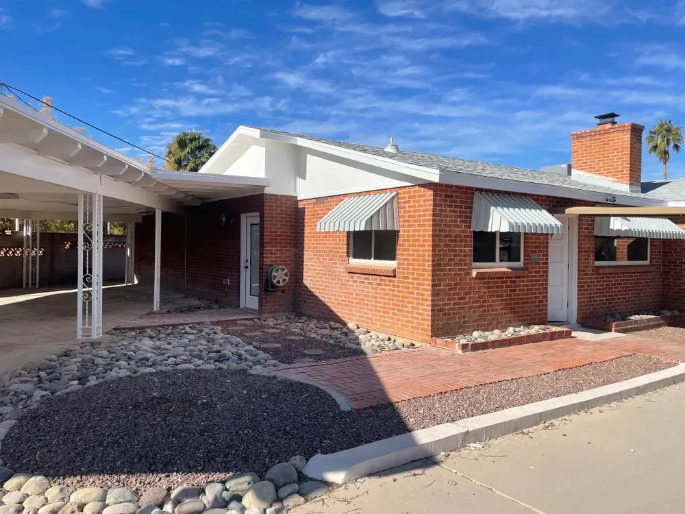 Brick house with a carport, awnings, and a brick pathway under a blue sky.