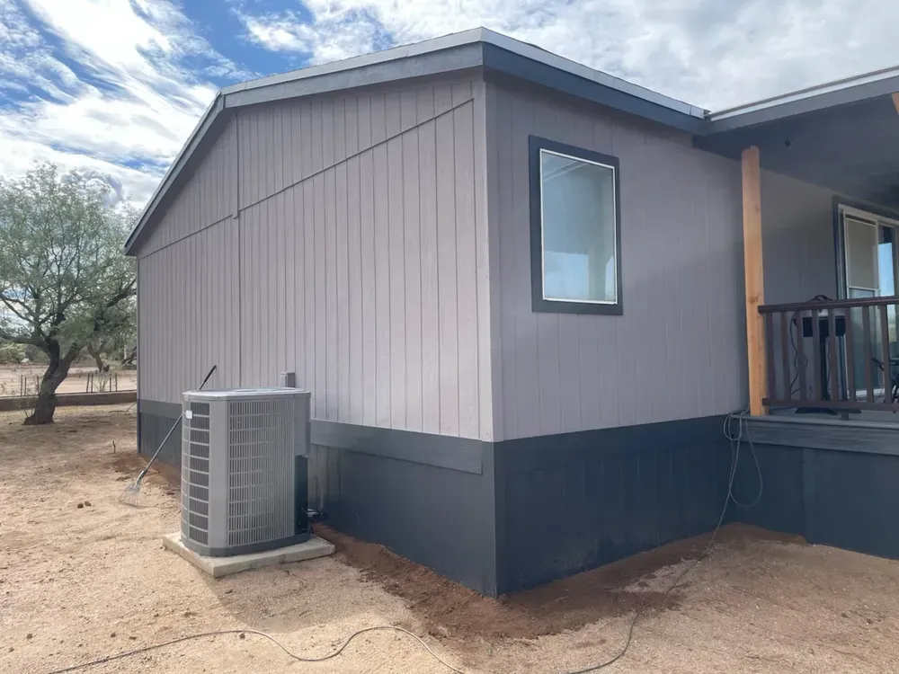 Exterior of a gray and black house with air conditioning unit. Cloudy sky and dry dirt ground.