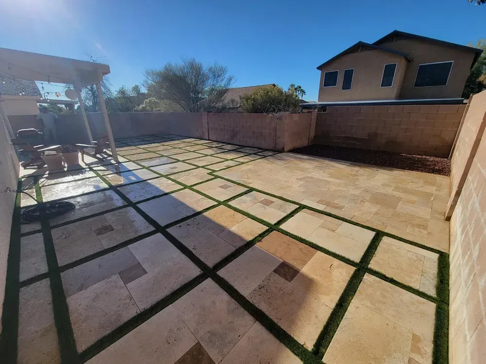 A backyard patio with tan stone tiles and green artificial grass strips, enclosed by a beige wall.