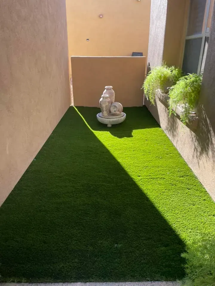 A small, enclosed patio with green artificial turf, a decorative fountain, and potted greenery.