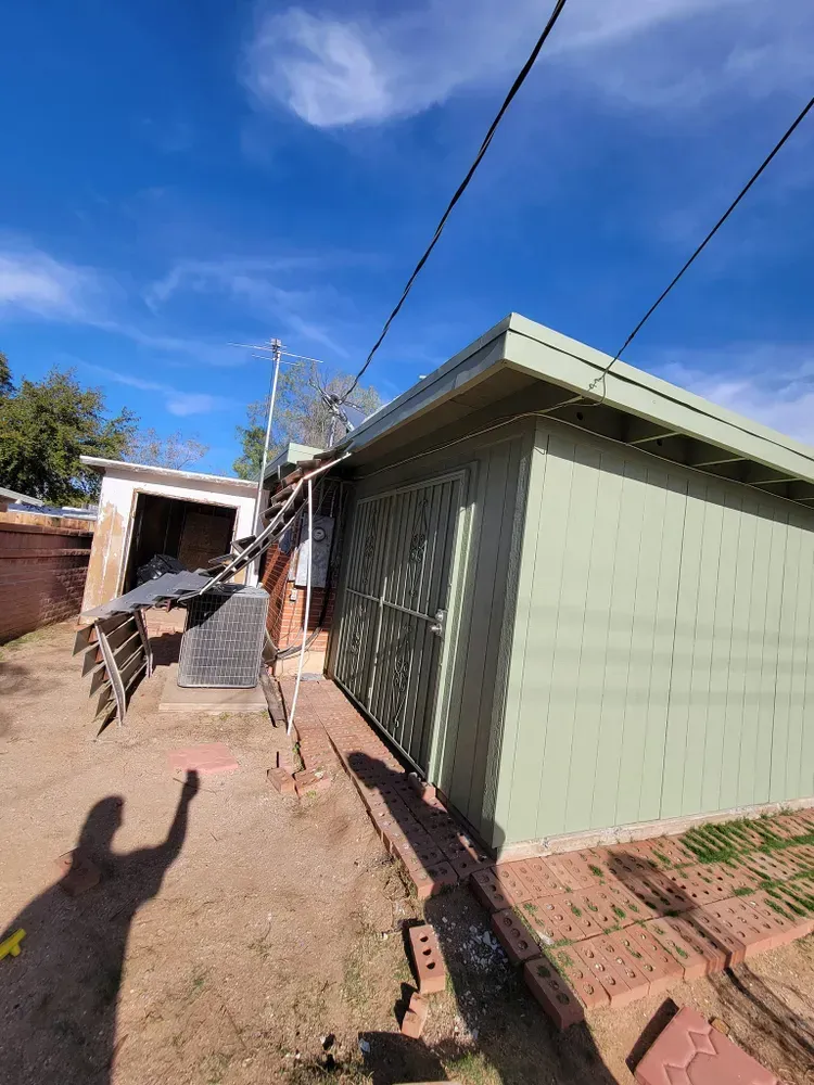 Green shed with metal door and overhead power lines. Sunny day, shadow of person in foreground.