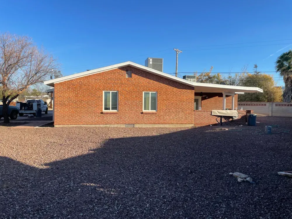 Brick house with two windows and a covered patio, in a gravel yard under a blue sky.