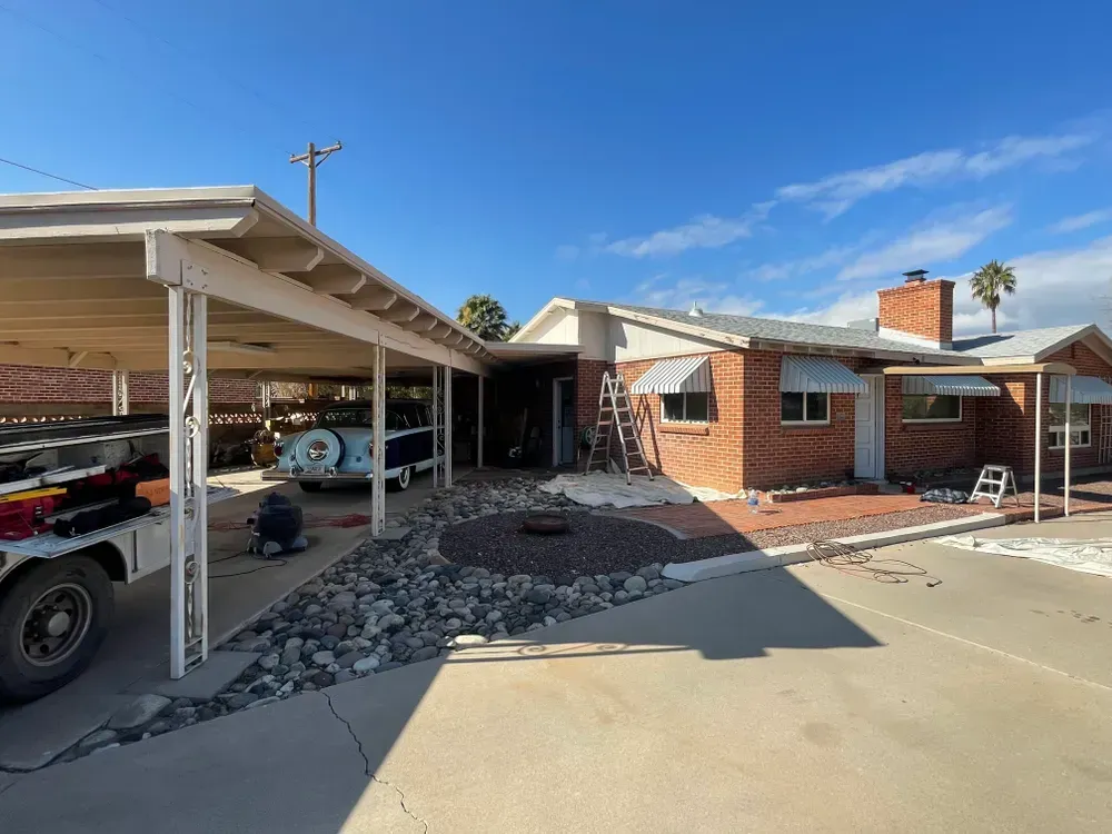 A brick house with a carport, undergoing construction. A vintage car is parked in the carport. Blue sky overhead.