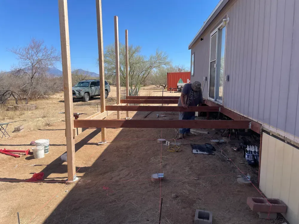 Man constructing a wooden deck outside a house in a desert setting; posts, beams, tools visible.