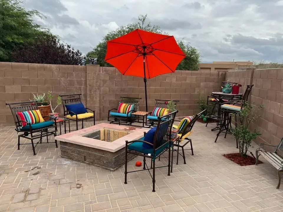 Patio with fire pit, seating, and orange umbrella against a block wall under cloudy skies.