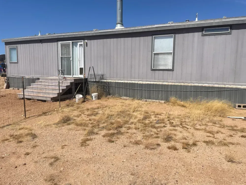 Gray mobile home with stairs, door, and windows. Dry, brown yard in front.
