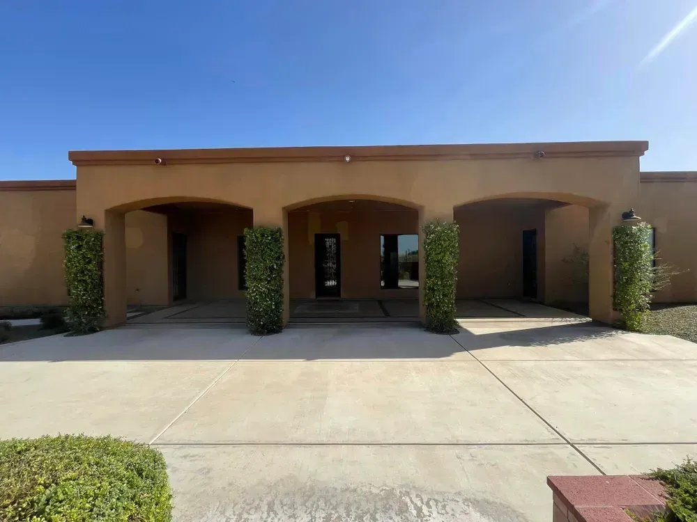 Tan building with three arched entryways, concrete path, and blue sky. Green shrubs flank the entry.