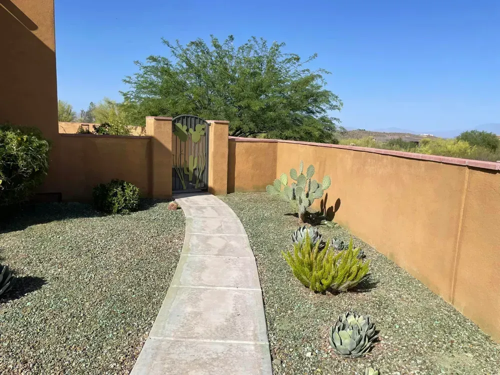 Stone path leading to a gate with a wall on either side, cactus, and shrubs with a blue sky.