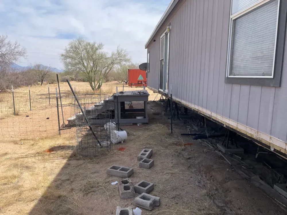 Mobile home exterior with cinder blocks and supports. Steps and desert landscape in the background.