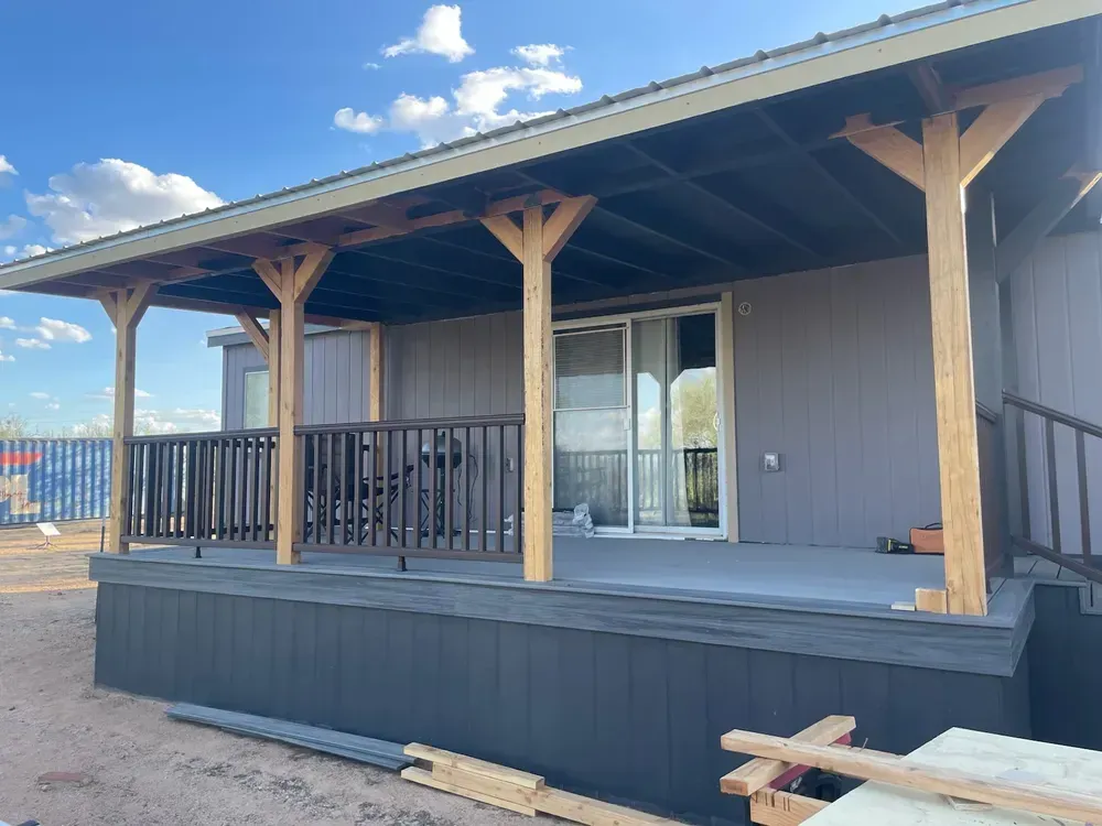 A house with a porch. Dark grey siding and deck, with light brown wooden supports. Blue sky.
