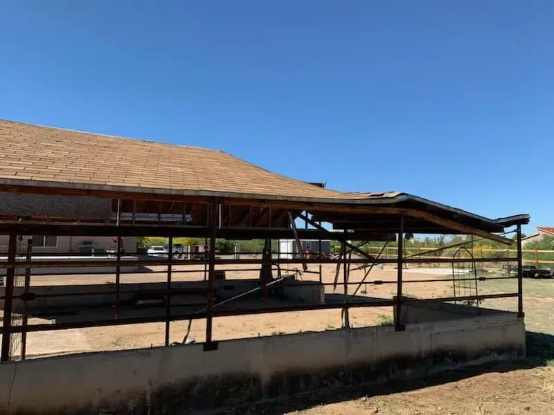 Dilapidated wooden structure with a damaged roof and metal fencing, under a blue sky.