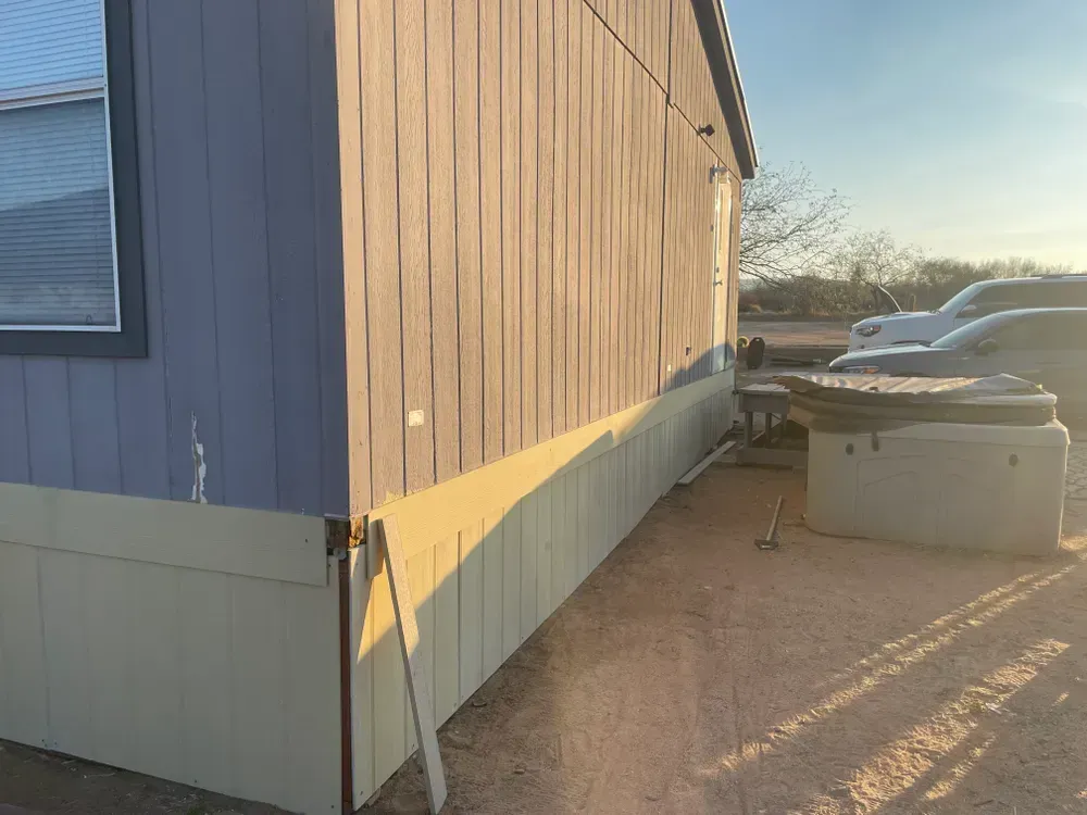 Exterior side view of a mobile home with blue siding, tan skirting, and a hot tub.