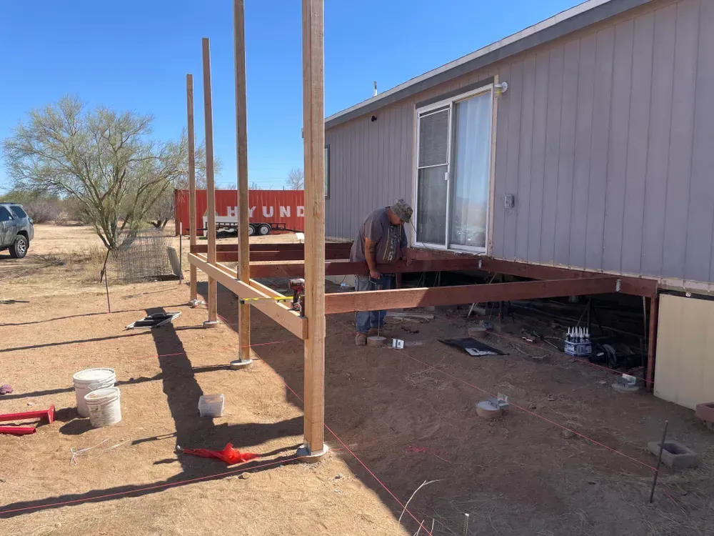 Man building a deck outside a house on a sunny day. Wooden posts, beams and deck under construction.