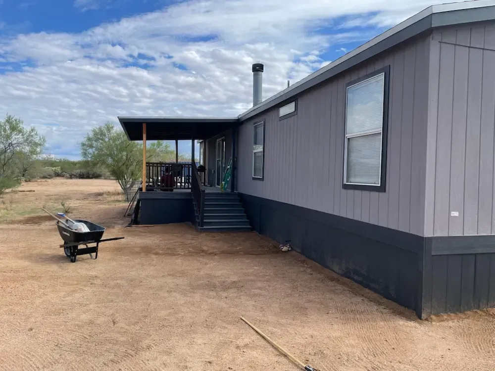 Gray mobile home with a porch, wheelbarrow, and dry, dirt yard under a cloudy blue sky.