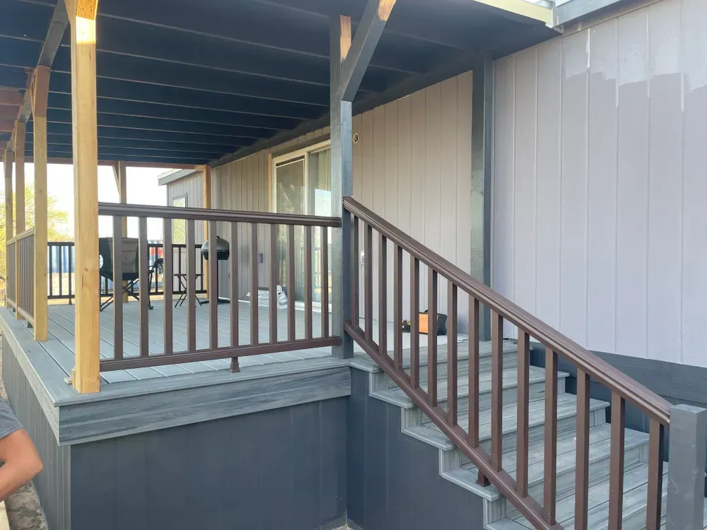 Covered porch and steps with brown railing and gray siding on a house.