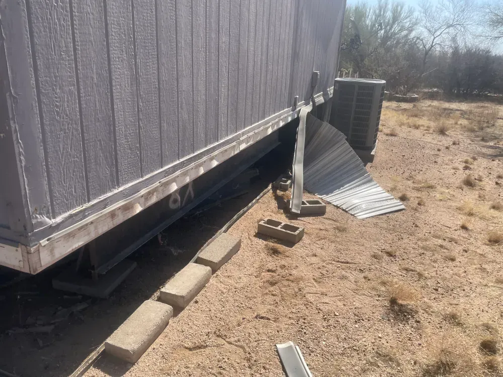 Side of a gray mobile home, with AC unit, concrete blocks, and damaged skirting in a dirt lot.