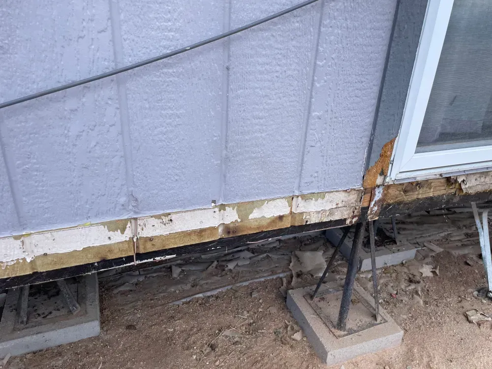 Damaged siding on a building with exposed wood and support blocks. Weathered exterior in a dirt setting.
