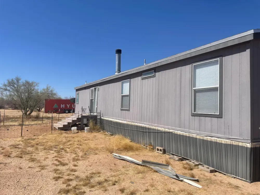 Gray mobile home with steps leading to a door, in a dry, grassy field under a blue sky.