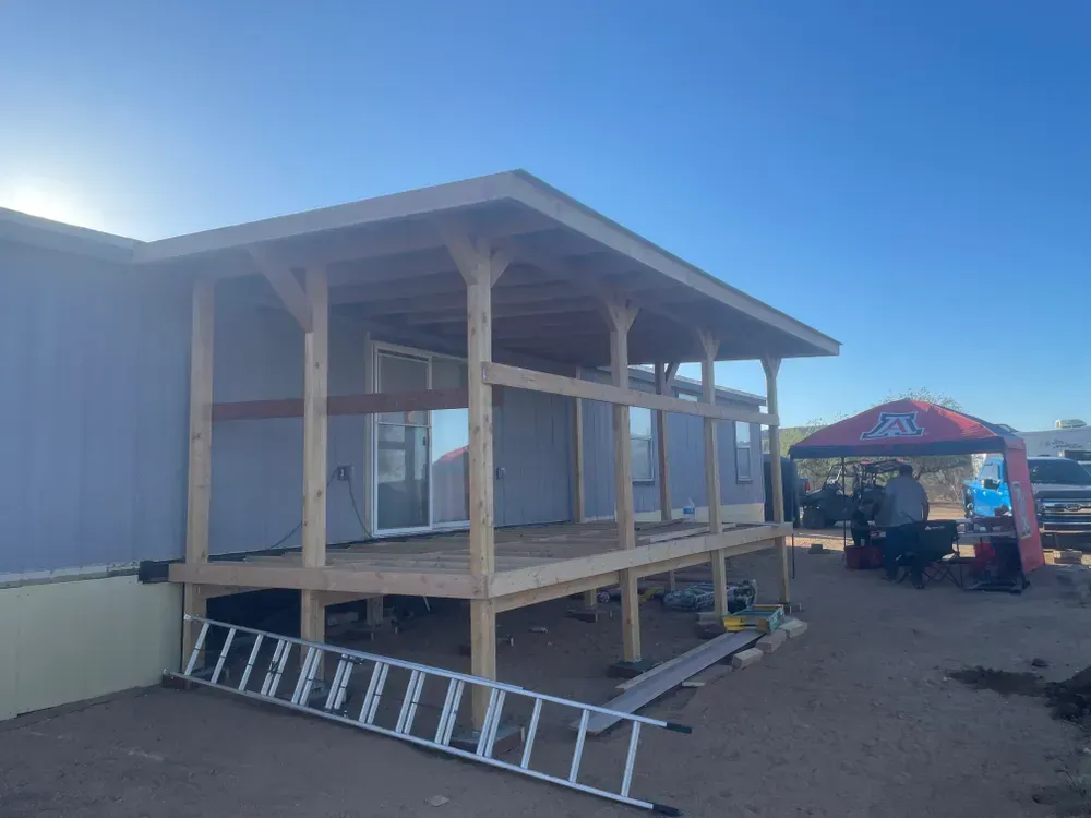 Porch under construction attached to a light blue house; wooden frame, ladder, workers, blue sky.