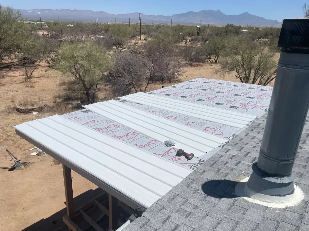 A metal roof being installed, with red underlayment visible, next to an existing shingled roof, outdoors.