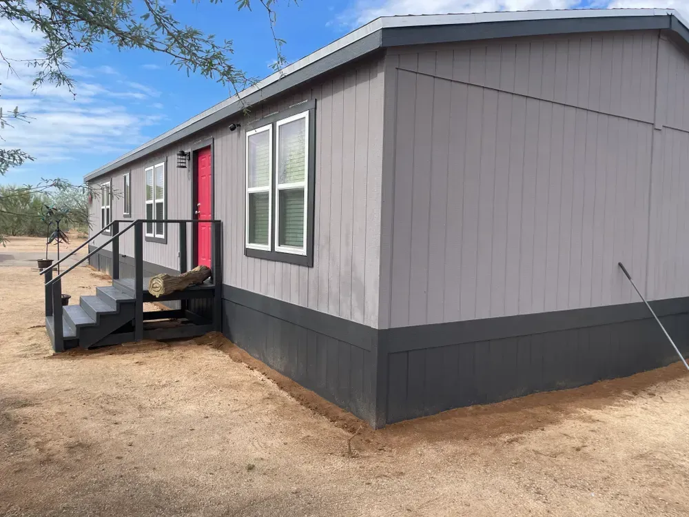 Gray mobile home with dark trim and red door, surrounded by dirt and sparse desert vegetation.