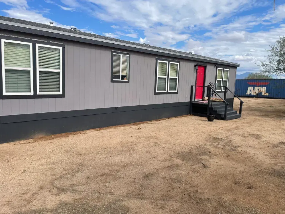 Gray mobile home with a red door, set in a sandy yard. A shipping container is in the background.