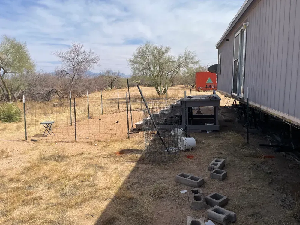 A chicken coop with a chicken in a dry, grassy yard beside a house; a small fence and distant hills.