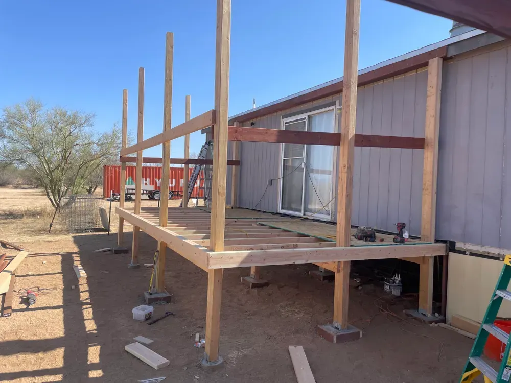 Deck construction in progress; wooden frame built alongside a light-colored building, outdoors on a sunny day.