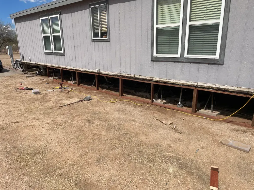 Mobile home with exposed foundation undergoing repair on dry, brown ground.